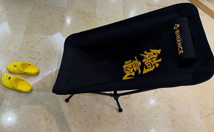 A black folding cot labeled 'F-BINANCE' with gold Japanese-style characters sits on a marble floor next to a pair of yellow Crocs shoes.