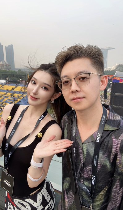 A young woman with long dark hair in a black top poses beside a bespectacled young man at what appears to be a Formula 1 event, both wearing lanyards featuring "OKX" branding. The caption humorously suggests the man appears so unfamiliar due to the woman's beauty filter that he jokes about mooching off her photo treatment for future appearances.
