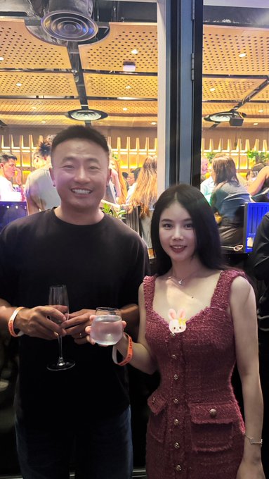 A man and woman posing together at what appears to be a restaurant, both holding drinks, with the woman wearing a pink textured dress.