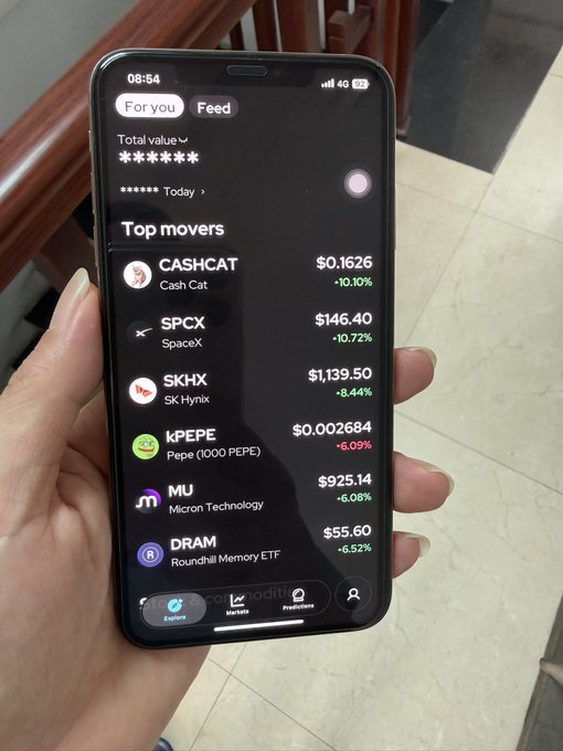 A photograph of a hand holding a smartphone displaying a trading app's 'Top movers' list, which shows CASHCAT ($0.1626, -10.10%), SPCX ($146.40, -10.72%), SKHX ($1,139.50, -8.44%), PEPE ($0.002684, -6.09%), MU ($925.14, +6.08%), and DRAM ($55.60, -6.52%).