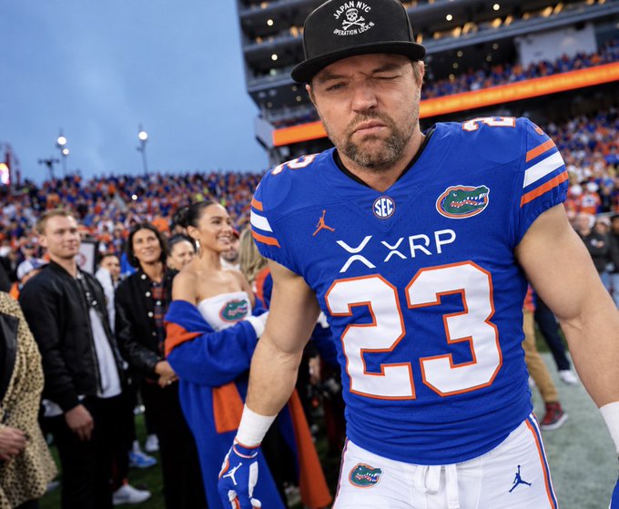 A Florida Gators football player wearing jersey number 23 walks off the field with a dejected expression, his blue jersey prominently displaying "XRP" branding across the chest along with the SEC and Gators logos, as a cheerleader and fans look on in the background. The image visually supports the post's claim about the XRP logo gaining mainstream visibility through its sponsorship placement on Florida Gators uniforms inside their stadium, "The Swamp."