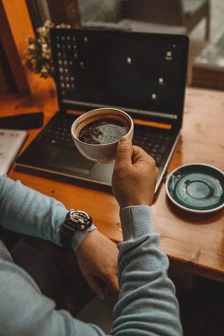 A person in a blue sweater holding a cup of coffee in front of an open laptop on a wooden table, wearing a black wristwatch.