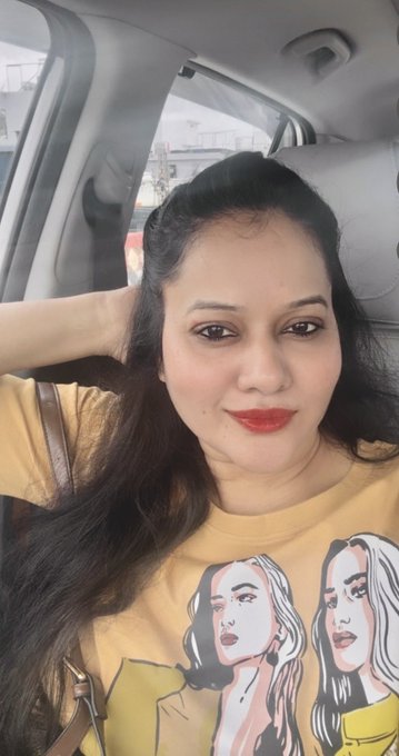 A woman with long dark hair and red lipstick takes a selfie inside a car, wearing a yellow graphic t-shirt featuring illustrated portraits of women.