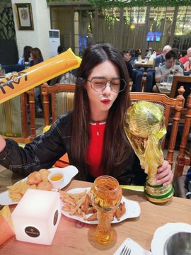 A woman holds a World Cup trophy and an inflatable noisemaker while sitting at a table with food and drinks, celebrating a win with Binance Traditional Chinese and predict.fun. The World Cup final viewing party and the winning of 4000 from @predictdotfun are directly related to the image.