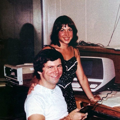 A vintage photograph of a smiling man and woman posing together indoors, with an older style television visible in the background.