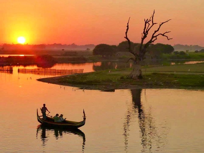 A serene landscape photo capturing a sunset over a river, featuring a long wooden boat carrying passengers and a rower, silhouetted against the golden sky, alongside a large, bare tree on the grassy bank.