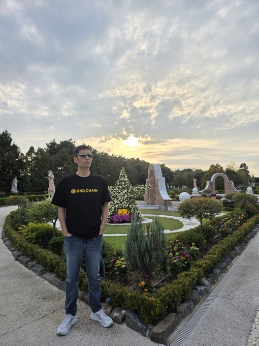 A young man wearing sunglasses, blue jeans, and a black t-shirt with the 'BNBCHAN' logo stands in a landscaped garden with flower beds and decorative archways at sunset.