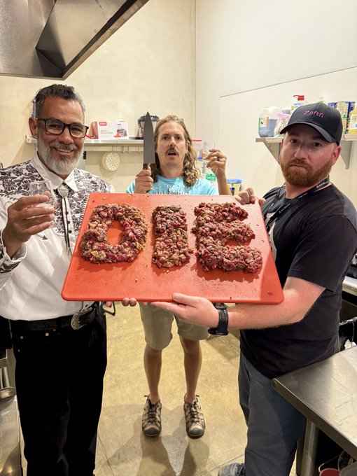 Three men in a kitchen proudly holding up a red cutting board with food items spelling 'DIE', with the man in the middle also holding a large knife.