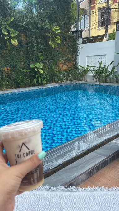 A first-person view of a hand holding an iced coffee from 'Ini Capuc' in front of a blue-tiled swimming pool surrounded by lush greenery.