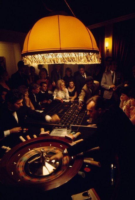 A vintage photograph shows people gathered around a casino gaming table, illuminated by a large fringed yellow lamp hanging above.