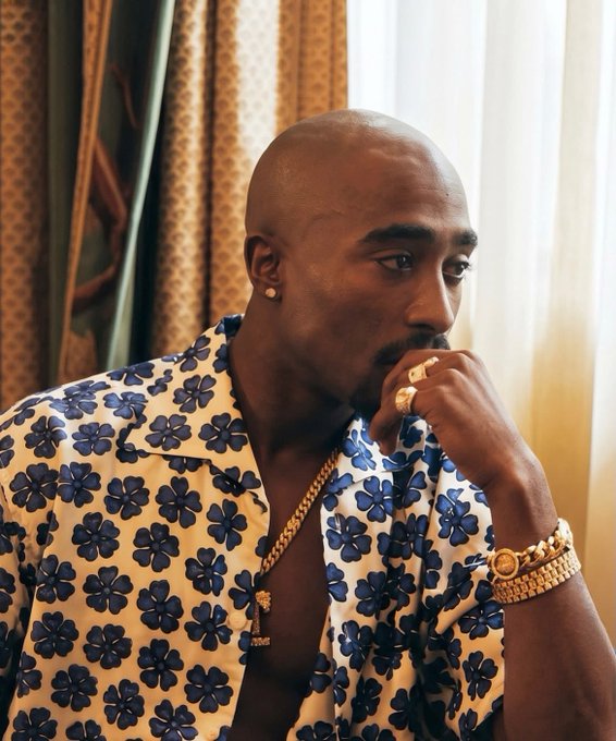 A man wearing a blue and white floral shirt, gold chain, rings, and a gold watch looks thoughtfully to the side.