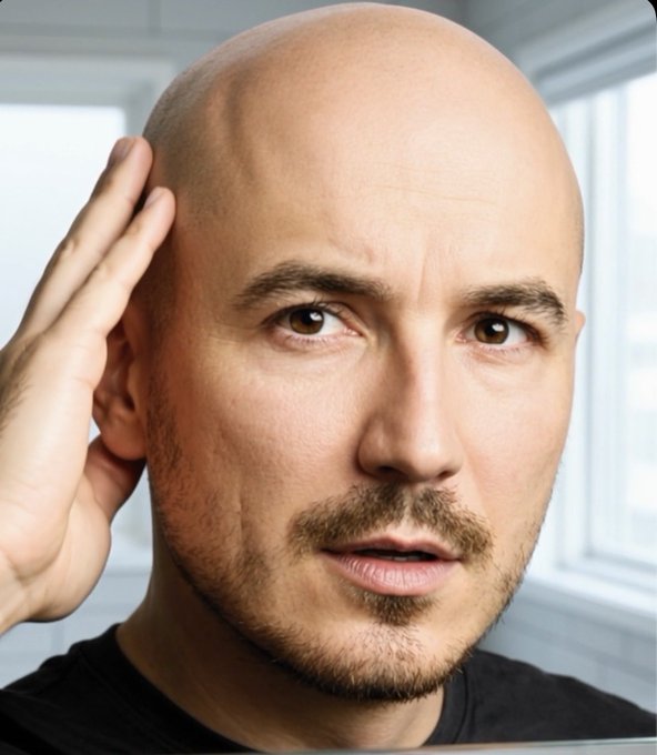 Close-up portrait of a bald man with a goatee, holding his hand to his head with a concerned expression, set against a blurred background of windows.