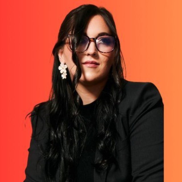 A portrait photo shows a woman with long dark hair, wearing tortoiseshell glasses, floral white earrings, and a black blazer, set against a bright red to orange gradient background.