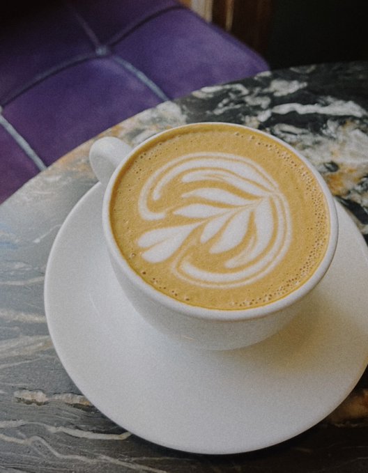A cup of coffee with intricate leaf-shaped latte art, served on a white saucer atop a marble-patterned table next to a purple tufted seat.