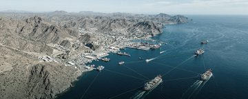 An aerial view of a busy harbor with cargo ships and cranes nestled among arid, rocky mountains along the coastline.