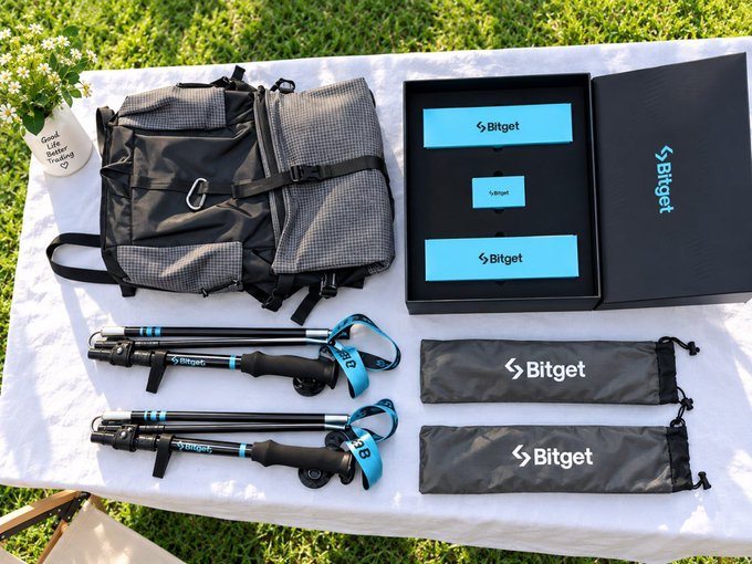 A Bitget promotional display on a table outdoors, featuring a black backpack, trekking poles, and pouches with the 'Bitget' logo.