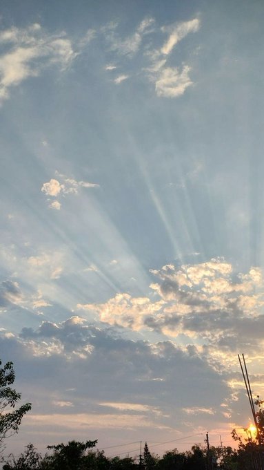 The image shows a beautiful sky with sun rays shining through clouds during sunrise or sunset. The sun appears to be setting, casting long shadows across the scene. In the background, there are trees that add depth and texture to the landscape. The photo is taken from an elevated position, providing a bird's eye view of the sky and its elements.