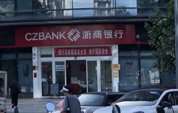 The image shows a street scene with a red bank building featuring a large sign that reads "CZBANK" written in Chinese characters. The bank's name is displayed in both English and Chinese, indicating its international presence. A person can be seen walking on the sidewalk next to the bank, while several cars are parked nearby. In addition, there is a motorcycle parked close to the building.