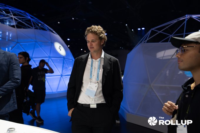 A man in a black suit stands at what appears to be a technology conference, with geometric dome structures visible in the background. The image has a 'The Rollup' watermark.