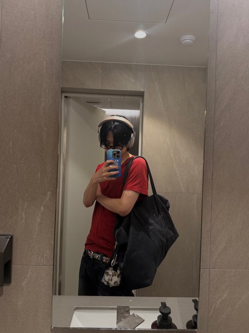 A mirror selfie showing a person wearing a red t-shirt and headphones, holding a blue phone while carrying a large black tote bag, taken in what appears to be a modern restroom or hotel bathroom with a sink visible in the foreground. The post hashtags "#LEO #리오" (LEO in Korean) likely reference VIXX's Leo, with the "260801" suggesting the date August 1, 2026.