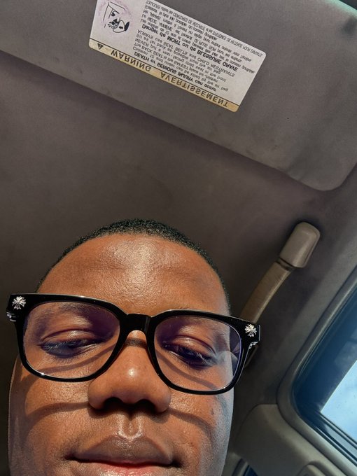 A close-up selfie of a man wearing glasses with floral details, looking down at the camera from inside a car, with an airbag warning label visible on the sun visor above him. The "gm winners." caption plays on the car setting, suggesting a good morning greeting while subtly referencing the vehicle's manufacturer, GM (General Motors).