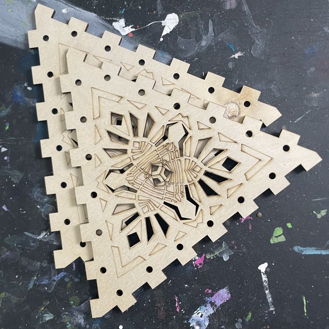An intricate, laser-cut wooden geometric piece rests on a dark, paint-splattered surface, featuring a complex central mandala-like pattern surrounded by a jagged border.