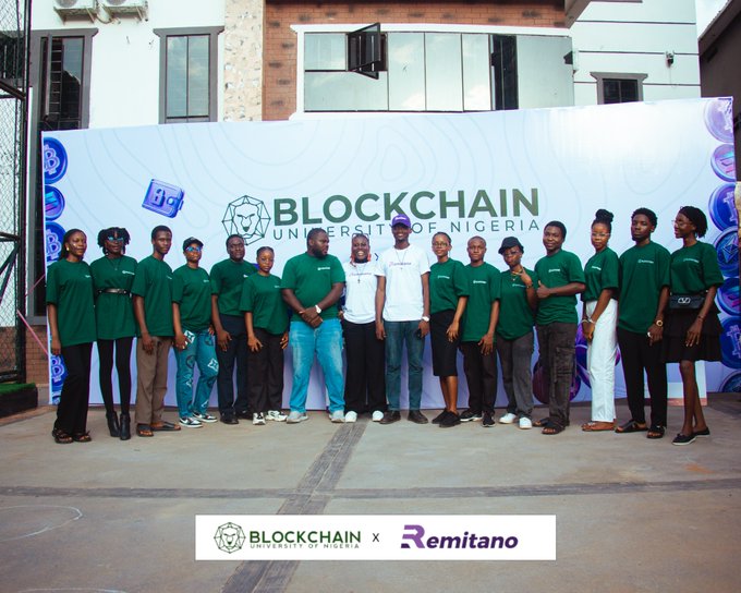 The image features a group of people standing together, posing for a photo with a banner that says "Blockchain" above them. The banner also has an email address written below it. There are several individuals in the group, all dressed similarly in green shirts. They appear to be gathered around a table or a display related to Bitcoin and Blockchain technology.