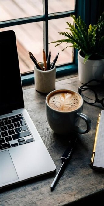 A cozy workspace featuring a laptop, a cappuccino with heart latte art, a cup of pencils, a potted plant, eyeglasses, a pen, and a notepad on a wooden desk by a window.