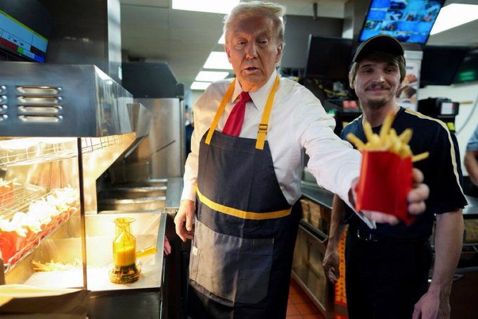 Donald Trump, wearing a white shirt, red tie, and McDonald's-themed apron, is pictured handing a carton of french fries to a McDonald's employee. The image humorously contrasts the high-stakes world of crypto trading with the mundane reality of fast-food work, suggesting a drastic shift in fortunes.
