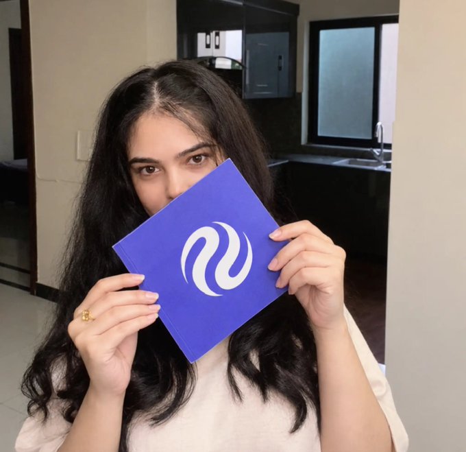 A woman with long dark hair holds a blue card featuring the white Injective logo in front of her face, relating to her post about discovering @injective through Zealy quests and becoming increasingly interested in $INJ and its ecosystem.