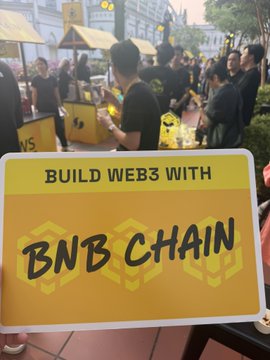A bold yellow sign reading "BUILD WEB3 WITH BNB CHAIN" is displayed prominently at the BNB Chain booth, capturing the event's builder-centric theme as crowds of attendees mingle in the background. The image directly ties to the post's observation about "builders" being BNB's unofficial mascot, highlighting why the $BUILDER token's sub-1M market cap seems undervalued given the community's pervasive "builder" branding.