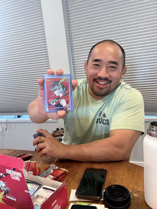 A smiling man in a green RVCA t-shirt sitting at a desk, proudly holding up a holographic anime-style trading card (appearing to be "Trace") with a Pokemon TCG backpack visible and a phone on the table.