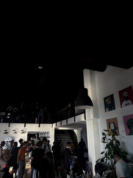 A lively crowd gathers at an artsy, dimly lit venue adorned with colorful framed portraits, where attendees mingle near a bar in what appears to be the Solana Summer House store. The post invites followers to meet up in person, and the bustling scene of people socializing and exploring the space reflects the energetic atmosphere of this exclusive crypto community event.