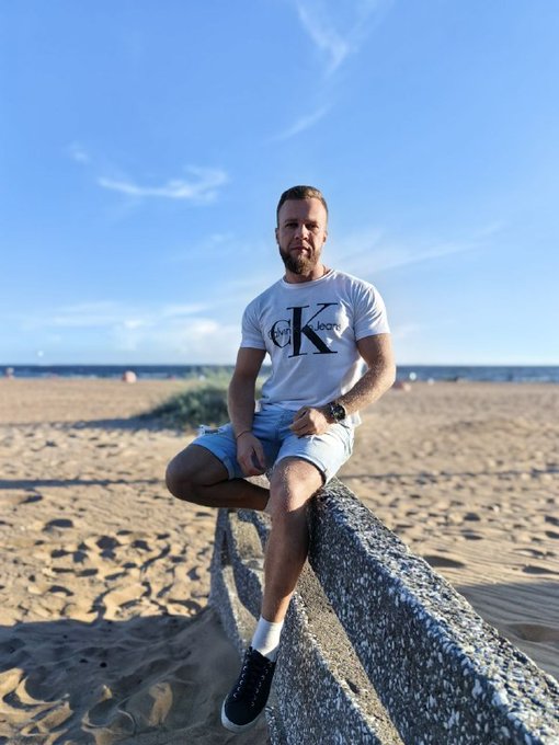 A man wearing a white Calvin Klein t-shirt, light blue shorts, and black sneakers sits on a stone barrier at a sandy beach under a clear blue sky.