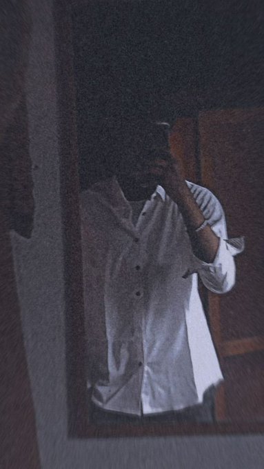 A dimly lit, slightly blurry mirror selfie shows a person wearing a white button-down shirt taking a photo with a smartphone, with their face obscured by the device.