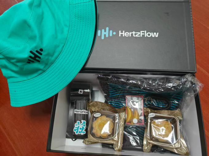 An opened black 'HertzFlow' branded gift box containing a teal cap, striped scarf, figurine, pins, and gold-wrapped snacks arranged on a wooden surface.