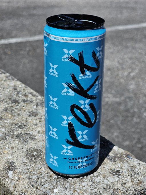 The image features a blue can of Keck Energy drinks placed on a concrete ledge or stone wall, with some text written on it that reads "KEK" and "world domination". The background appears to be an outdoor setting, possibly a street or sidewalk area.