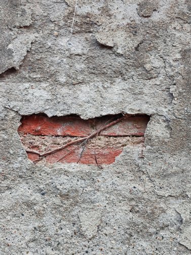 A weathered concrete wall with a hole revealing red brick underneath, with exposed wires or roots running through the opening.