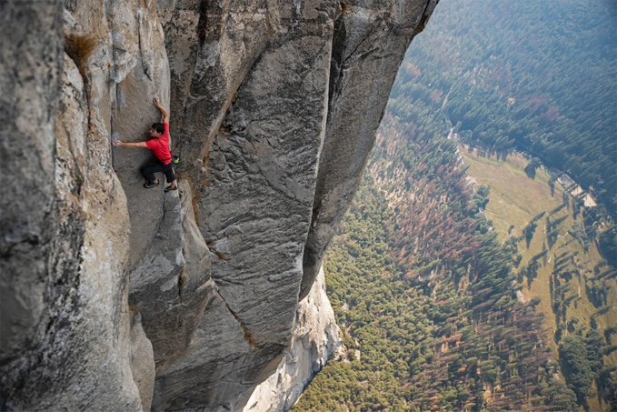 A lone climber in a red shirt scales a sheer granite cliff face high above a forested valley, embodying the precarious and focused effort of buying at the market bottom. The image visually captures the risk, isolation, and required nerve of entering a position when others are too afraid to hold on.