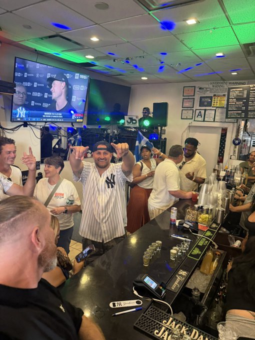 A lively bar scene with patrons watching a Yankees baseball game on TV; a man in a Yankees jersey and cap gestures enthusiastically with both hands raised.