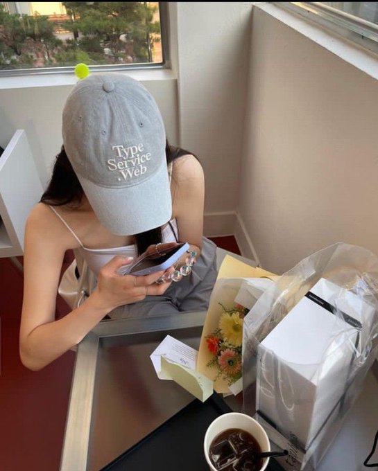 A person wearing a gray "Type Service Wep" cap sits at a table looking down at their phone, surrounded by a bouquet of flowers, a wrapped gift, and a cup of iced coffee. The image contrasts with the post's tech-focused message about AI taking over markets, juxtaposing a quiet, personal moment of stillness with the urgency of "AI is building" and the race to adopt AI trading tools.