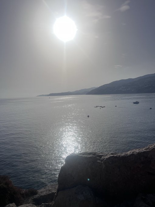 A serene seascape with the bright sun shining over calm water, mountains in the distance, and small boats scattered across the bay, viewed from a rocky shoreline.