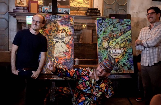 Three men pose in front of two colorful, graffiti-style paintings, with the central seated man wearing a patterned shirt and traditional cap making hand gestures.