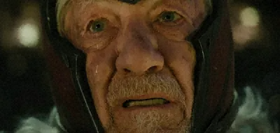 A close-up of an older man with a white beard wearing a hood, gazing upward with a determined expression.
