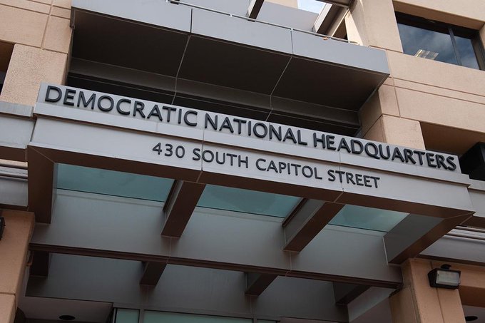 A low-angle shot of the entrance to the Democratic National Headquarters at 430 South Capitol Street, featuring bold black lettering above the doorway. The image underscores the post's claim about the DNC's financial troubles following DOGE budget cuts, highlighting the party's central Washington headquarters.
