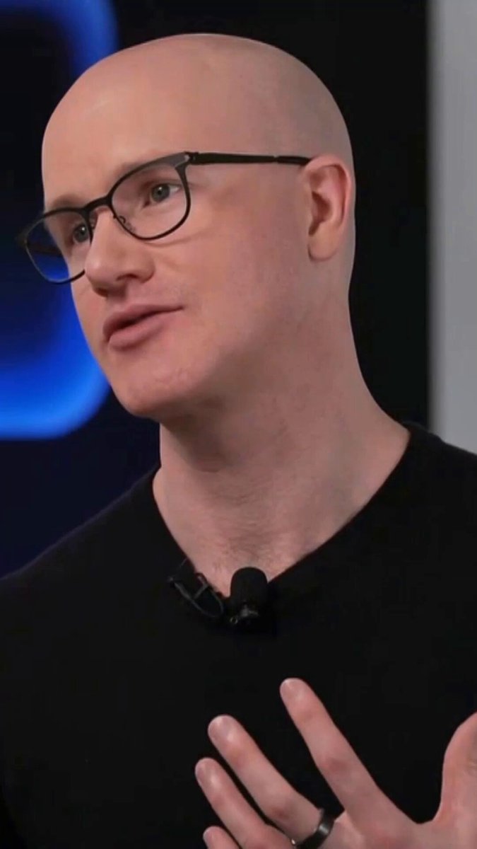 The image features a bald man wearing glasses, giving him a serious expression as he speaks into a microphone. The background of the image is blurred, drawing attention to the man with the microphone. There are no other objects or people visible in the image, making the focus on the man and his words the primary subject matter.