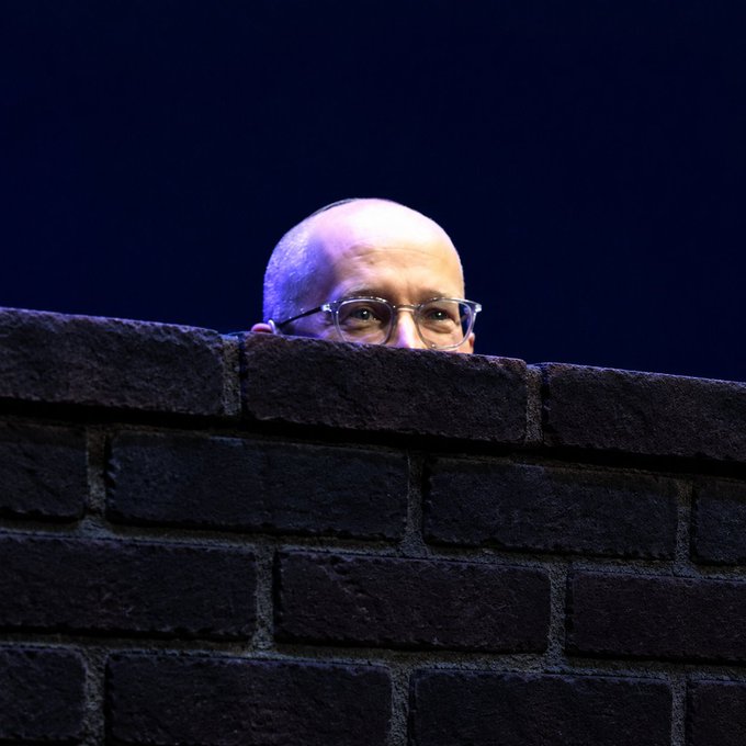 A bald man wearing glasses peers over the top of a dark brick wall against a dark blue background.