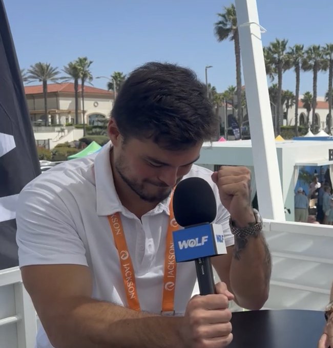 A man with a beard being interviewed on camera, holding a 'WOLF' branded microphone while wearing a white polo shirt and a Jackson lanyard, with palm trees visible in the background.