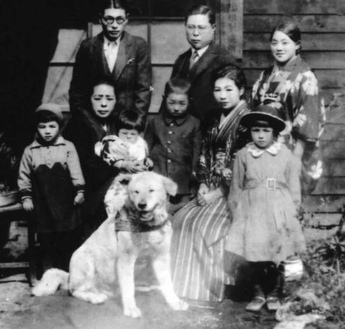 A vintage black-and-white family portrait featuring men in suits, women in kimonos, and children gathered around a large light-colored dog, likely an Akita, in front of a wooden house.