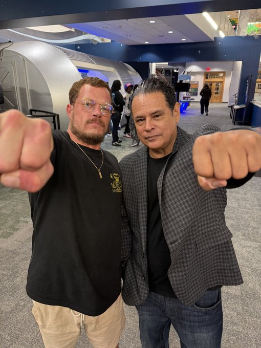 Two men posing with their fists extended toward the camera in a playful manner, standing in what appears to be a space or science museum with a space capsule in the background.